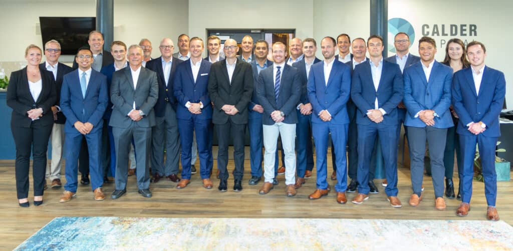 A professional group photo of a diverse team of 25 individuals in business attire, standing in two rows in an office setting. They are smiling and looking towards the camera.